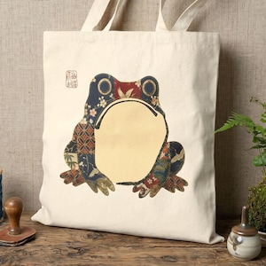 Puede incluir: Bolso tote de color crema con un diseño de rana grande. La rana está compuesta por varias telas estampadas en tonos azules, rojos y dorados. Un pequeño sello rojo es visible en la parte superior izquierda. El bolso tiene dos asas color crema.