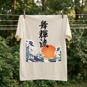 A cream-colored t-shirt hangs on a clothesline outdoors with wooden clothespins against a blurred forest backdrop. The ukiyo-e-inspired t-shirt is decorated with a vibrant design of an orange sun and tumultuous blue waves. Black Japanese kanji script meaning "Infinite Flow" is printed vertically.