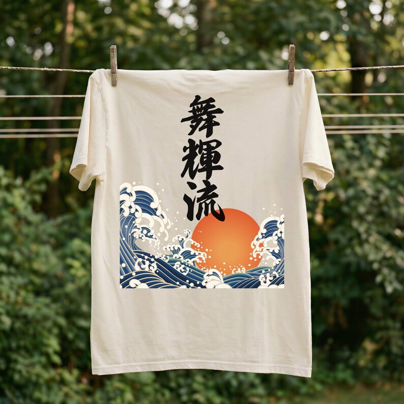 A cream-colored t-shirt hangs on a clothesline outdoors with wooden clothespins against a blurred forest backdrop. The ukiyo-e-inspired t-shirt is decorated with a vibrant design of an orange sun and tumultuous blue waves. Black Japanese kanji script meaning "Infinite Flow" is printed vertically.