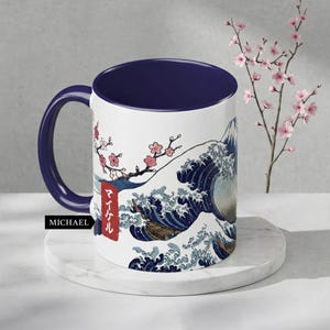 May include: White ceramic mug with a navy blue interior and handle. The mug features a Japanese wave design with a mountain in the background, cherry blossoms, and Japanese text. The word "MICHAEL" is printed on the side.