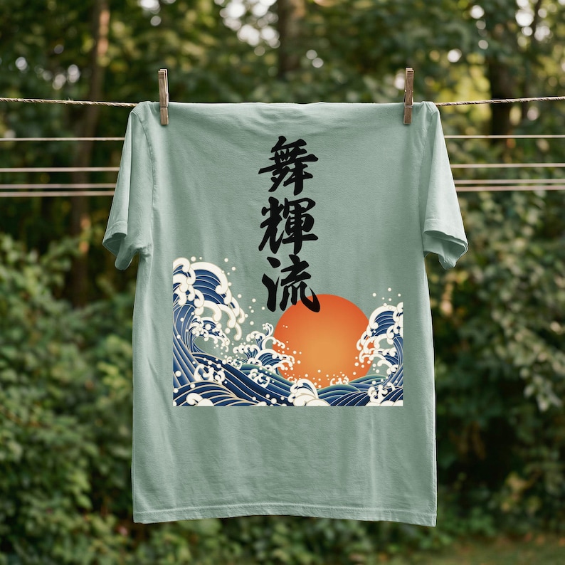 A light green / bay colored t-shirt hangs on a clothesline outdoors with wooden clothespins. A forest is blurred in the background. The t-shirt features a dynamic design in the ukiyo-e style of a large orange sun and dramatic blue waves. Black Japanese kanji script meaning "Infinite Flow" is printed vertically.