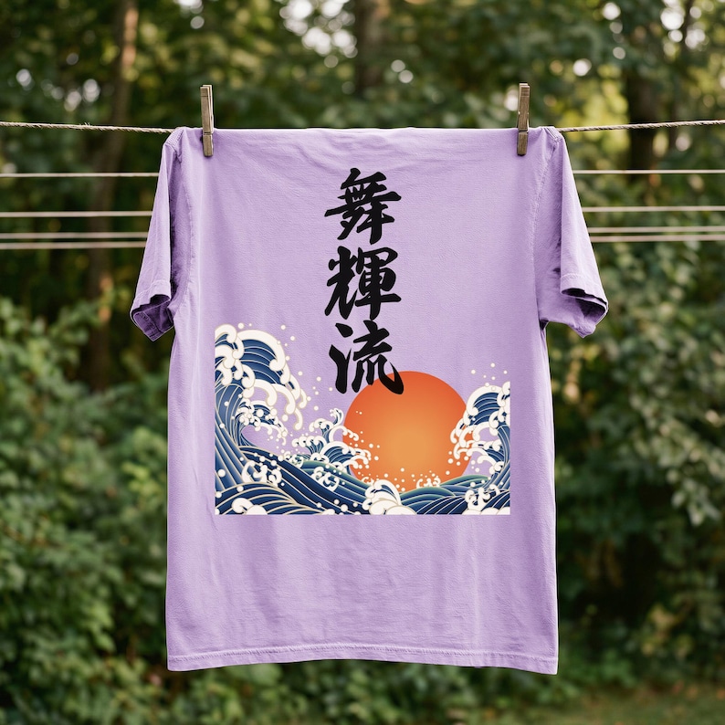 A purple / orchid colored t-shirt hangs on a clothesline outdoors with wooden clothespins against a background of blurred trees and foliage. The vibrant ukiyo-e style design on the t-shirt features a glowing orange sun and dynamic blue waves. Black Japanese kanji script meaning "Infinite Flow" is printed vertically.