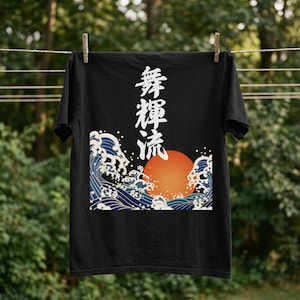 A black t-shirt hangs on a clothesline outdoors with wooden clothespins. A forest is blurred in the background. The dramatic ukiyo-e style design on the t-shirt features a glowing orange sun and tumultuous blue waves. Black Japanese kanji script meaning "Infinite Flow" is printed vertically.