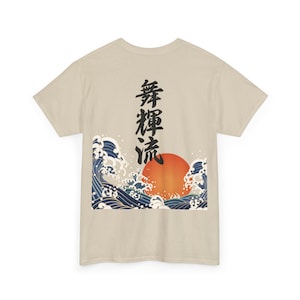 The back view of a light beige or cream-colored t-shirt laid flat against a white background. The shirt features a large, detailed graphic print in the lower-center section, depicting vertical black Japanese Kanji characters rising above a traditional ukiyo-e style design of a large orange sun and crashing blue ocean waves.