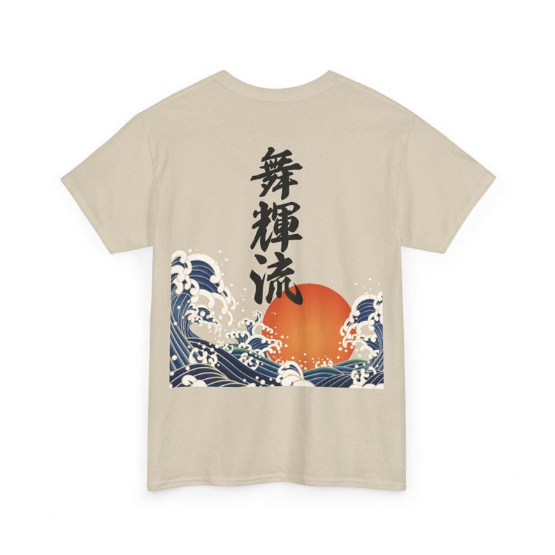 The back view of a light beige or cream-colored t-shirt laid flat against a white background. The shirt features a large, detailed graphic print in the lower-center section, depicting vertical black Japanese Kanji characters rising above a traditional ukiyo-e style design of a large orange sun and crashing blue ocean waves.