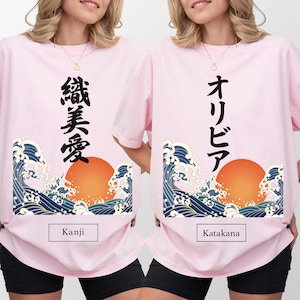 Two women wearing light pink oversized t-shirts showcasing different Japanese script styles. The shirt on the left features vertical Kanji characters, labeled "Kanji" below. The shirt on the right features vertical Katakana characters, labeled "Katakana" below. Both shirts are decorated with a traditional Japanese ukiyo-e style artwork of a large orange sun and blue ocean waves.