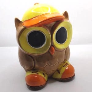 May include: A ceramic owl ornament with large yellow eyes and a yellow cap with an orange brim. The owl is brown with orange and yellow accents on its feet. The figurine has a whimsical, cartoonish design.