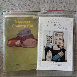May include: Two craft pattern booklets in plastic sleeves. The left booklet, Sleeping Country Cat, shows three fabric cats and states 15 inches long. The right booklet, Keeping You In Stitches Cat Talk, displays various stitched cat-themed items.