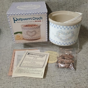 May include: A Rival Potpourri Crock electric simmering cooker with a white and blue checkered pattern and a floral heart design. The set includes the original box, a white stoneware crock with a handle, instruction manual, and a bag of potpourri.