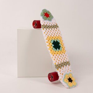 Peut inclure: Un skateboard blanc avec une housse crochetée colorée. La housse présente un motif floral avec des accents jaunes, verts et bleus. Le skateboard a des roues rouges.