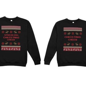 以下が含まれることがあります： 黒い長袖スウェットシャツ2枚。それぞれに、赤色の「SPREADING CHRISTMAS CHEER」と「CHRISTMAS CHEER」の文字、トナカイ、雪の結晶、クリスマスツリーが描かれたお祝いのデザインが施されています。