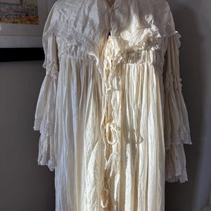 May include: Cream-colored vintage nightgown with a high collar, lace trim, and long, ruffled sleeves. The gown is displayed on a dress form, showcasing its flowing fabric and delicate details. The nightgown has a tie closure.