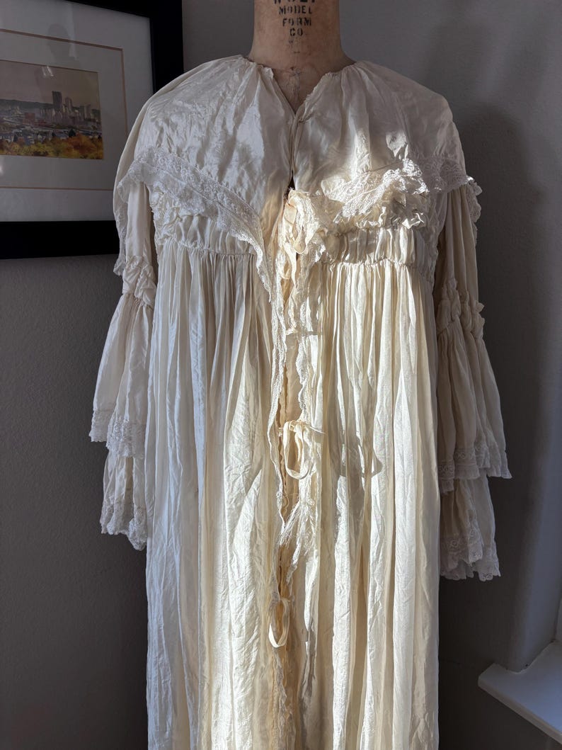 May include: Cream-colored vintage nightgown with a high collar, lace trim, and long, ruffled sleeves. The gown is displayed on a dress form, showcasing its flowing fabric and delicate details. The nightgown has a tie closure.