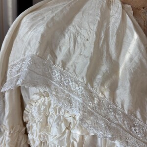 May include: Ivory-colored vintage dress with a ruffled bodice and skirt. The dress features a delicate lace trim along the neckline and sleeves. The fabric has a subtle sheen, suggesting a silk or satin material. The dress is displayed on a mannequin.
