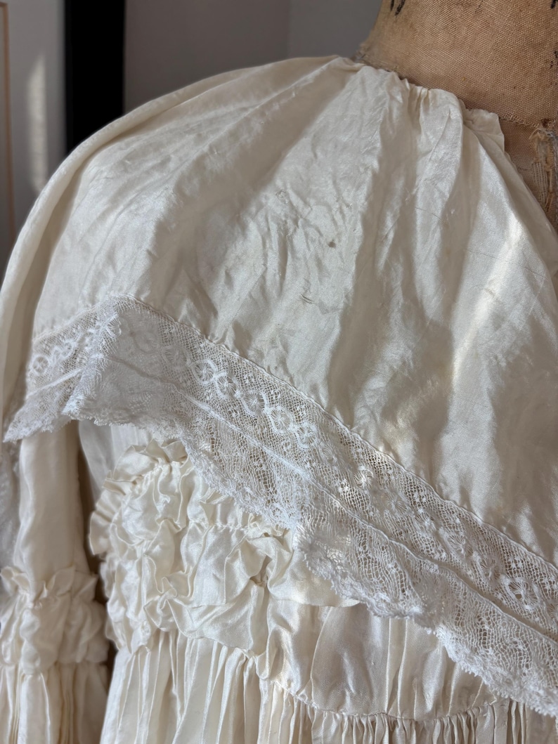 May include: Ivory-colored vintage dress with a ruffled bodice and skirt. The dress features a delicate lace trim along the neckline and sleeves. The fabric has a subtle sheen, suggesting a silk or satin material. The dress is displayed on a mannequin.