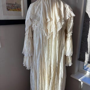 May include: Cream-colored vintage dress with long sleeves and ruffled detailing. The dress is displayed on a mannequin, showcasing its intricate lace and flowing fabric. The dress has a high neckline and a floor-length hemline.