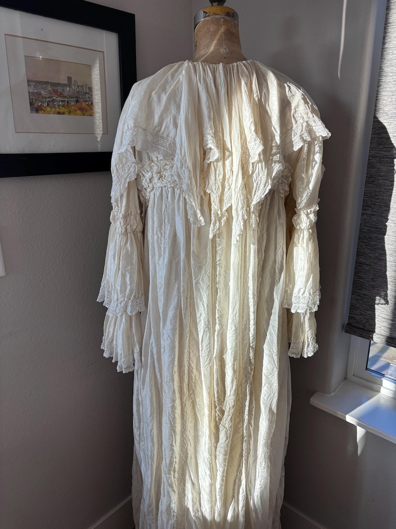 May include: Cream-colored vintage dress with long sleeves and ruffled detailing. The dress is displayed on a mannequin, showcasing its intricate lace and flowing fabric. The dress has a high neckline and a floor-length hemline.