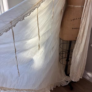 May include: Off-white vintage dressing gown with lace trim and ribbon ties. The gown is draped over a vintage dress form. The fabric has a soft, flowing appearance, with a ruffled hemline. The dress form has the words "COLLAR MODEL" printed on it.
