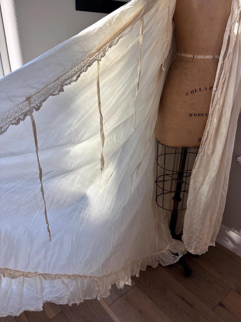 May include: Off-white vintage dressing gown with lace trim and ribbon ties. The gown is draped over a vintage dress form. The fabric has a soft, flowing appearance, with a ruffled hemline. The dress form has the words "COLLAR MODEL" printed on it.
