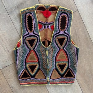 May include: A crocheted vest with a geometric pattern in shades of brown, green, purple, and yellow. The vest has a suede patch on the back and a button closure.