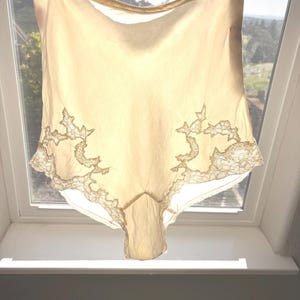 May include: A pair of vintage high-waisted underwear in a pale yellow hue, hanging in front of a window. The underwear features delicate lace detailing along the bottom edges, adding a touch of elegance to the garment.
