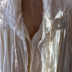 May include: Cream-colored, vintage-style blouse with a deep V-neck. The blouse features delicate lace trim along the collar and sleeves, and flowing fabric with a slightly wrinkled texture.