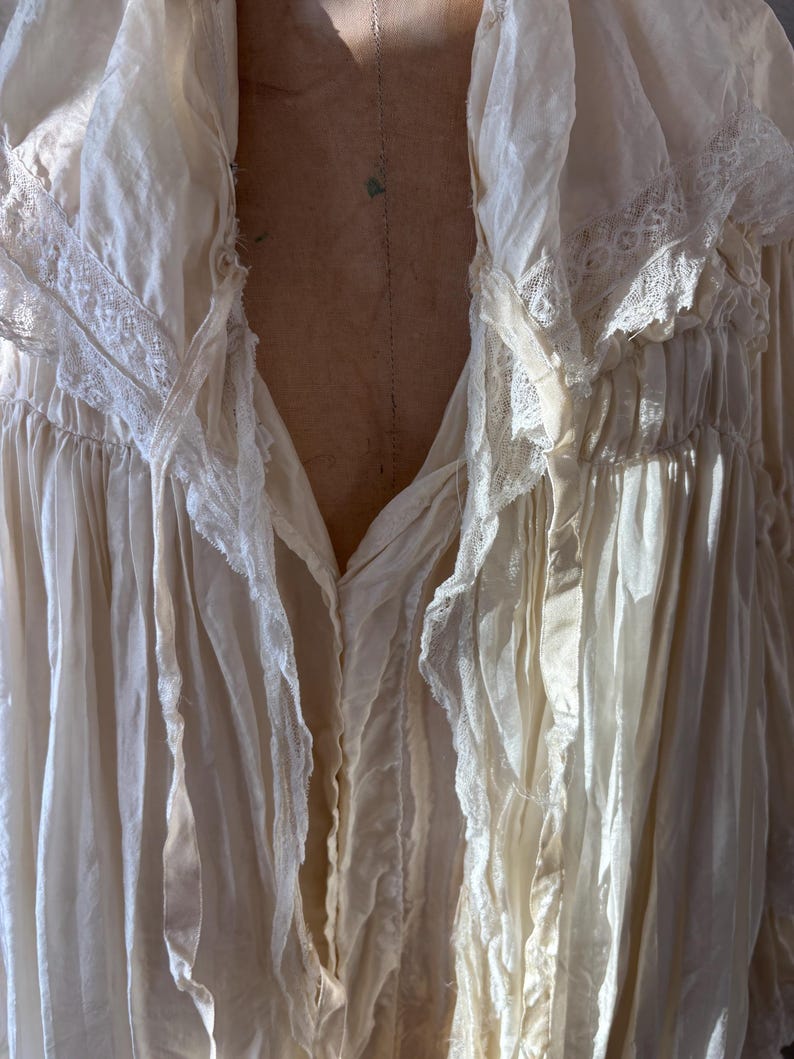 May include: Cream-colored, vintage-style blouse with a deep V-neck. The blouse features delicate lace trim along the collar and sleeves, and flowing fabric with a slightly wrinkled texture.