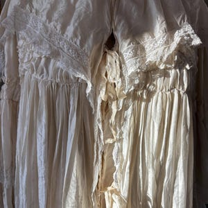 May include: Cream-colored vintage gown with lace trim and ruffled details. The long-sleeved garment features a V-neckline and a flowing, textured fabric. The dress appears to be made of a delicate material, possibly silk or a similar fabric.