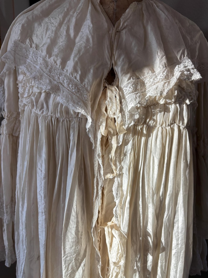 May include: Cream-colored vintage gown with lace trim and ruffled details. The long-sleeved garment features a V-neckline and a flowing, textured fabric. The dress appears to be made of a delicate material, possibly silk or a similar fabric.