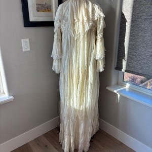 May include: A cream-colored, vintage-style dress with long, ruffled sleeves and a tiered skirt. The dress is displayed on a mannequin, showcasing its intricate lace and textured fabric. The dress is floor-length and appears to be made of a lightweight material.