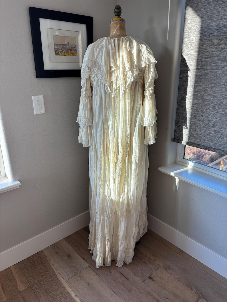 May include: A cream-colored, vintage-style dress with long, ruffled sleeves and a tiered skirt. The dress is displayed on a mannequin, showcasing its intricate lace and textured fabric. The dress is floor-length and appears to be made of a lightweight material.