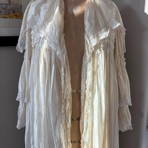 May include: Cream-colored vintage robe with lace trim and ruffled sleeves. The long, flowing garment is displayed on a vintage dress form. The robe has a delicate, romantic aesthetic, with a soft, textured appearance.