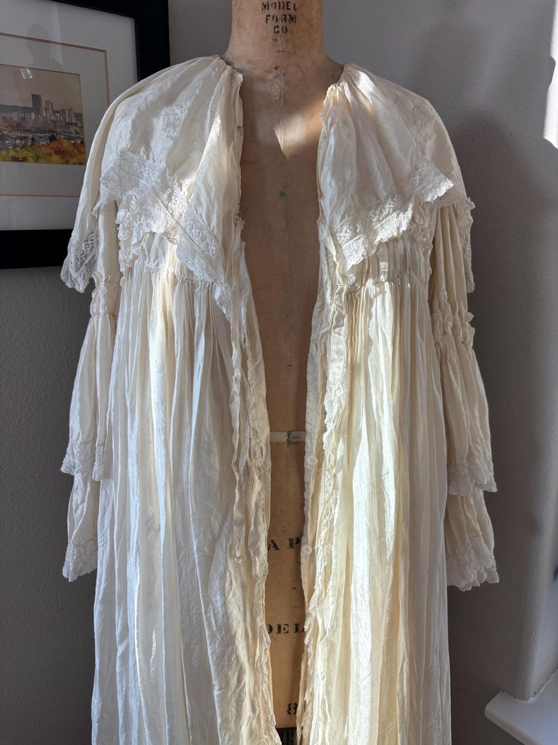 May include: Cream-colored vintage robe with lace trim and ruffled sleeves. The long, flowing garment is displayed on a vintage dress form. The robe has a delicate, romantic aesthetic, with a soft, textured appearance.