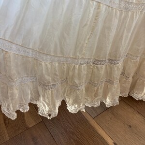 May include: A close-up of a cream-colored, layered fabric with delicate lace trim. The fabric appears to be a skirt or dress, with multiple tiers and a flowing, lightweight texture. The lace adds a vintage, romantic touch.