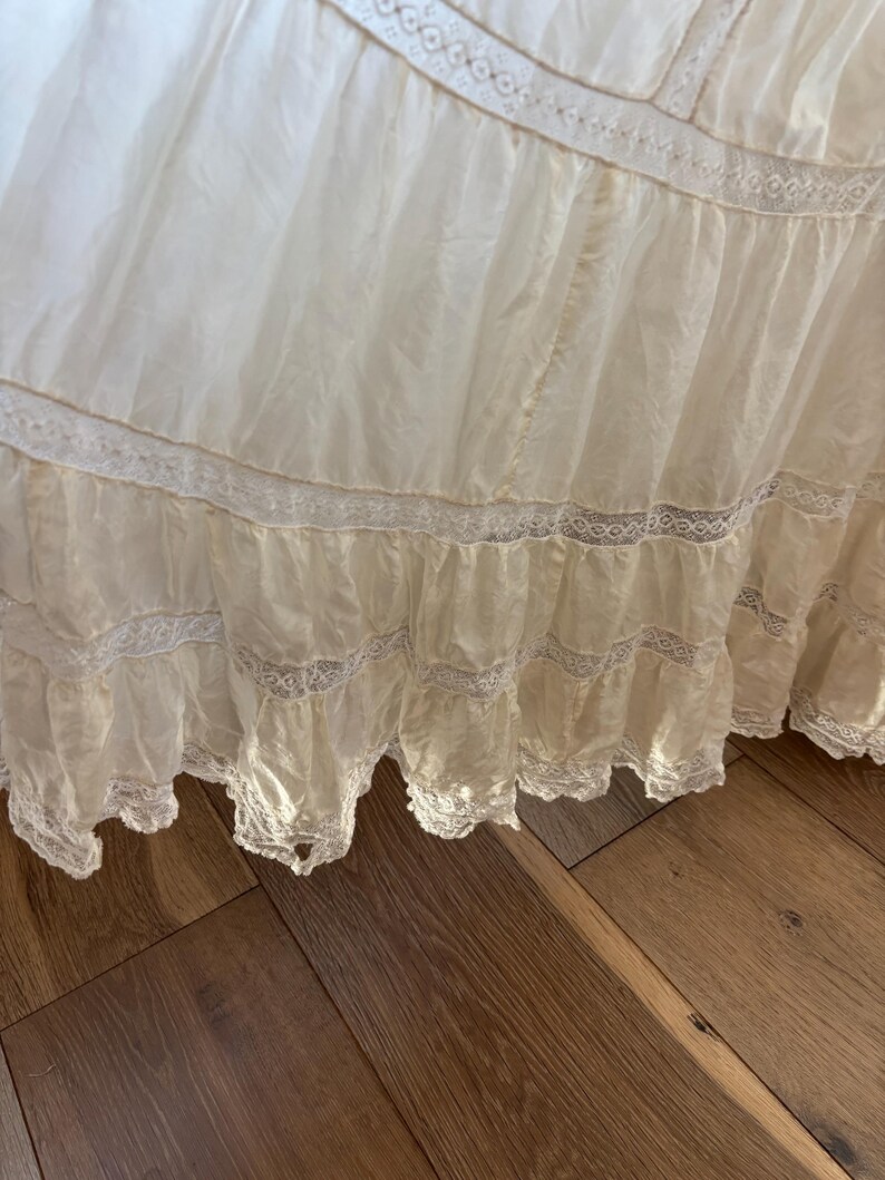 May include: A close-up of a cream-colored, layered fabric with delicate lace trim. The fabric appears to be a skirt or dress, with multiple tiers and a flowing, lightweight texture. The lace adds a vintage, romantic touch.