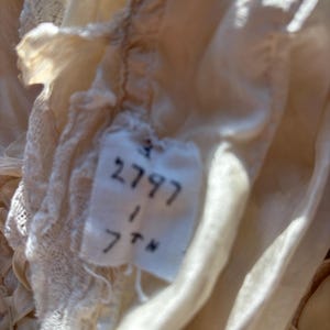May include: Close-up of a vintage cream-colored fabric with delicate lace trim. A small tag is visible, displaying the numbers "2797", "1", and "7th" in black ink. The fabric appears to be silk or a similar delicate material.