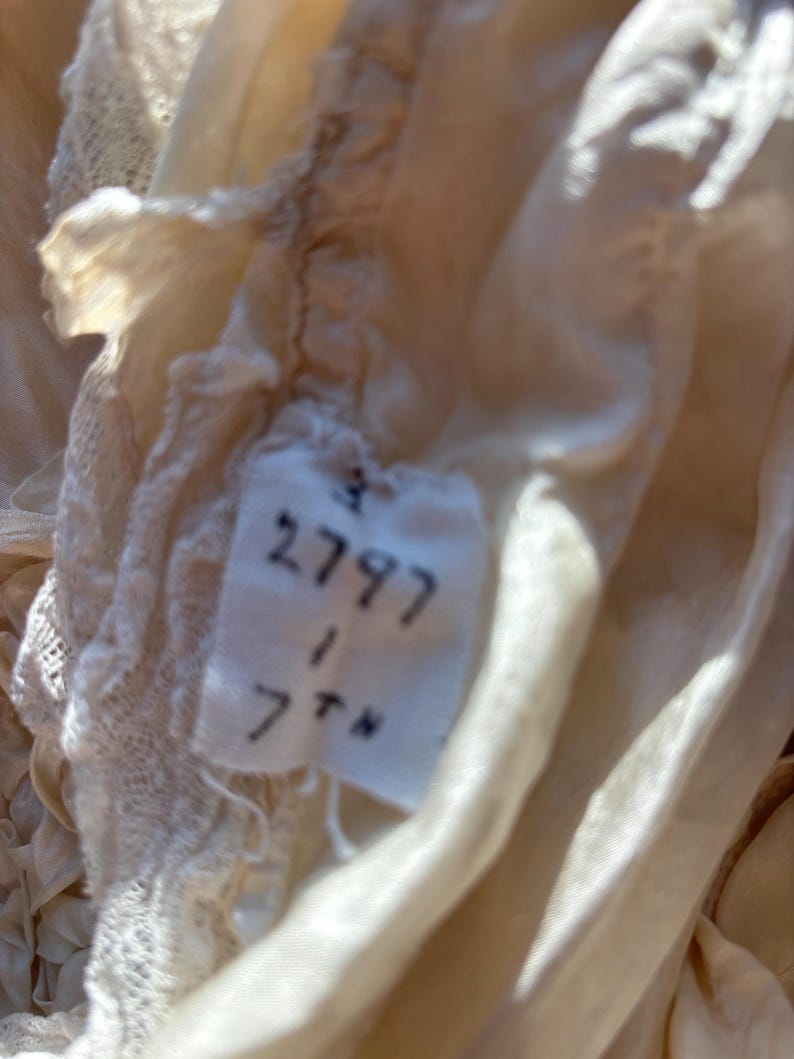May include: Close-up of a vintage cream-colored fabric with delicate lace trim. A small tag is visible, displaying the numbers "2797", "1", and "7th" in black ink. The fabric appears to be silk or a similar delicate material.