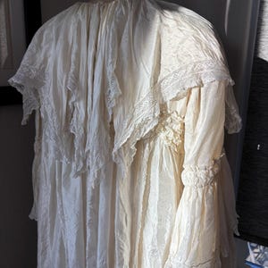 May include: Cream-colored vintage dress with a high neckline and long sleeves. The dress features delicate lace trim on the collar, sleeves, and bodice. The fabric appears to be a lightweight material, with a flowing, layered design.
