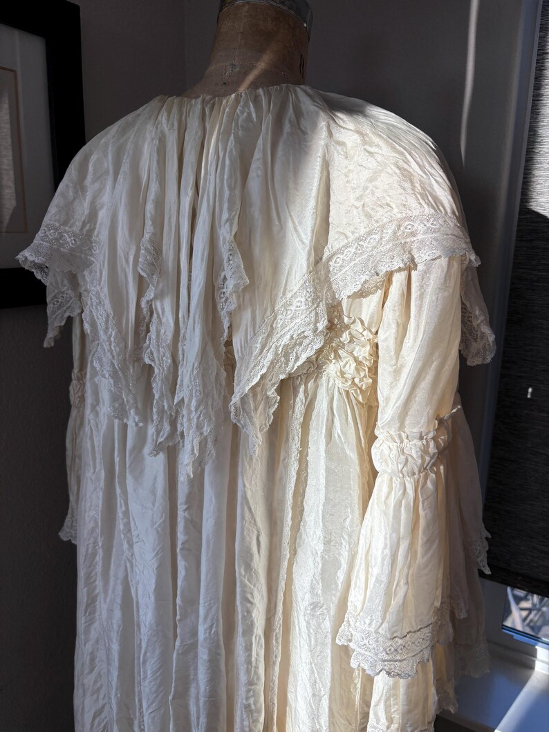 May include: Cream-colored vintage dress with a high neckline and long sleeves. The dress features delicate lace trim on the collar, sleeves, and bodice. The fabric appears to be a lightweight material, with a flowing, layered design.