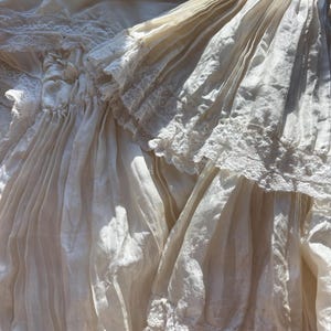 May include: Close-up of antique ivory-colored fabric with delicate lace trim. The fabric is draped and folded, creating a textured effect. The lace adds a touch of elegance to the vintage textile. The image captures the details of the aged material.