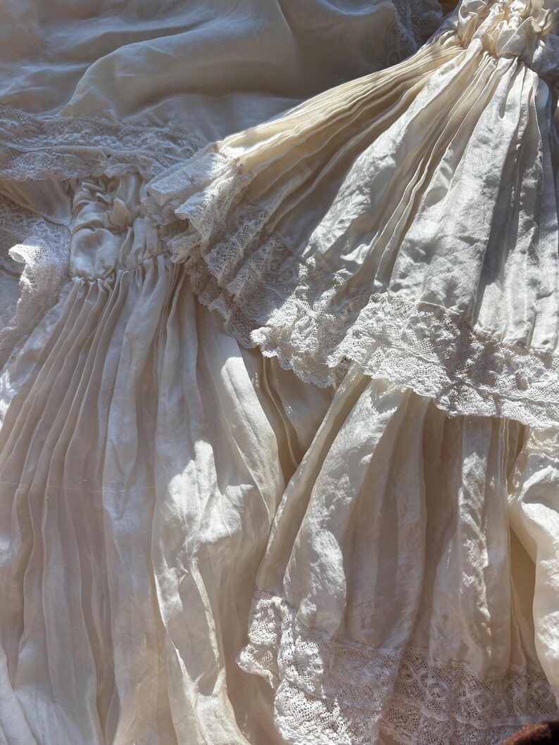 May include: Close-up of antique ivory-colored fabric with delicate lace trim. The fabric is draped and folded, creating a textured effect. The lace adds a touch of elegance to the vintage textile. The image captures the details of the aged material.