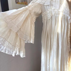 May include: Cream-colored vintage dress with long, ruffled sleeves and delicate lace trim. The dress features a high neckline and a flowing silhouette, suggesting a romantic or historical style. The fabric appears lightweight and slightly sheer.