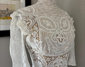 May include: A white high-neck blouse featuring intricate lace panels, floral embroidery, and sheer fabric sections. The garment has long sleeves with lace detailing and a fitted bodice constructed from a combination of delicate lace and cotton fabric.