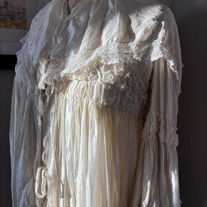 May include: Cream-colored vintage dress with a high ruffled collar and lace trim. The dress features long, flowing sleeves with ruffled cuffs and a gathered bodice. The fabric appears to be lightweight and slightly sheer, with a soft, textured appearance.