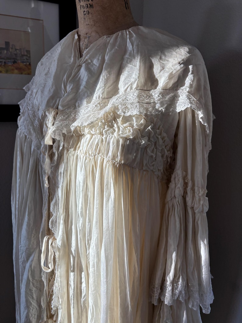 May include: Cream-colored vintage dress with a high ruffled collar and lace trim. The dress features long, flowing sleeves with ruffled cuffs and a gathered bodice. The fabric appears to be lightweight and slightly sheer, with a soft, textured appearance.