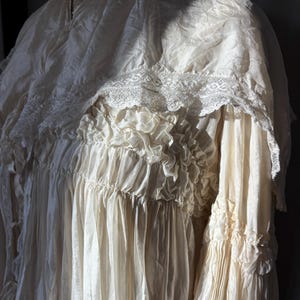 May include: An antique cream-colored dress with a high neckline and intricate lace detailing. The dress features layers of ruffles and pleats, creating a flowing, textured appearance. The sleeves are long and gathered at the cuffs, adding to the vintage aesthetic.