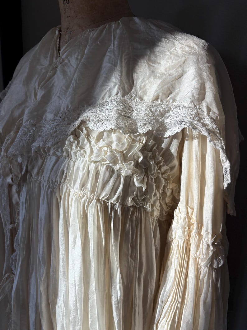 May include: An antique cream-colored dress with a high neckline and intricate lace detailing. The dress features layers of ruffles and pleats, creating a flowing, textured appearance. The sleeves are long and gathered at the cuffs, adding to the vintage aesthetic.