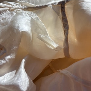 May include: Close-up of a cream-colored, semi-sheer fabric with a delicate texture. The fabric appears to be layered, with visible seams and a subtle sheen. The image captures the folds and draping of the material, highlighting its lightweight quality.