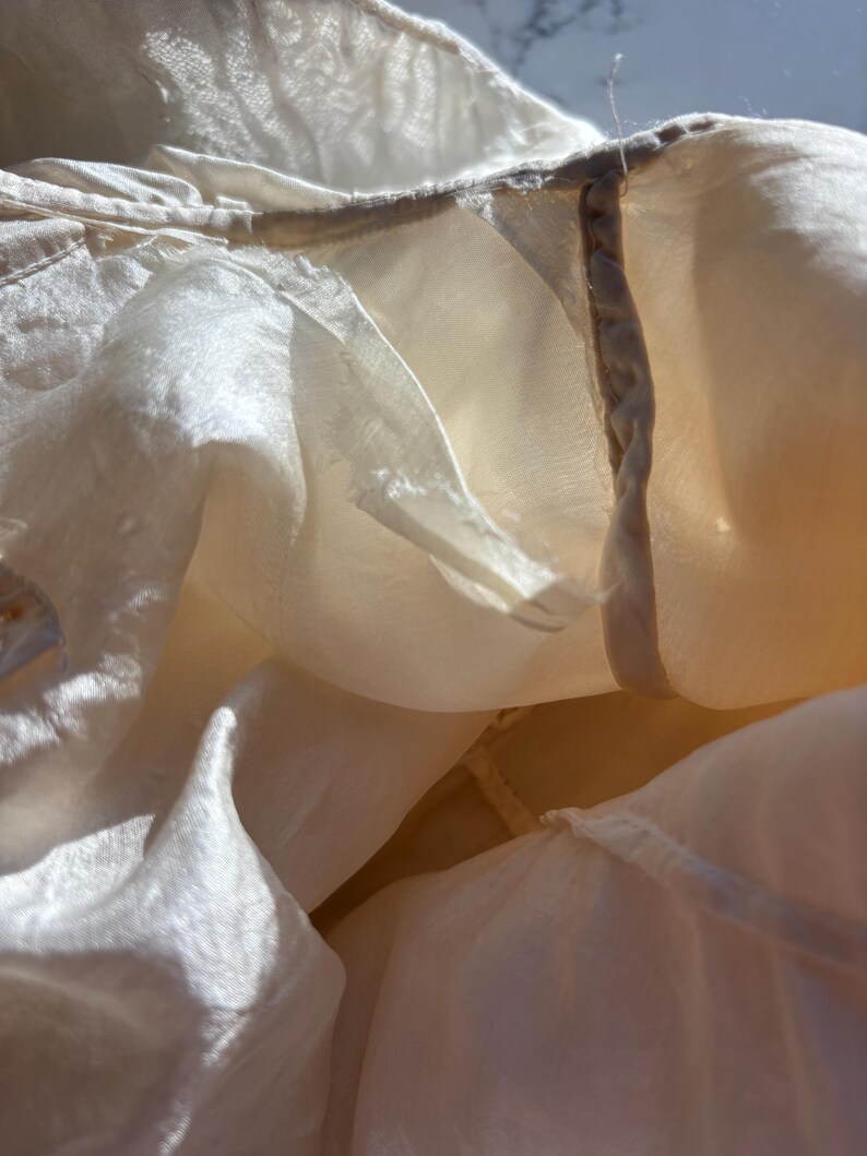 May include: Close-up of a cream-colored, semi-sheer fabric with a delicate texture. The fabric appears to be layered, with visible seams and a subtle sheen. The image captures the folds and draping of the material, highlighting its lightweight quality.