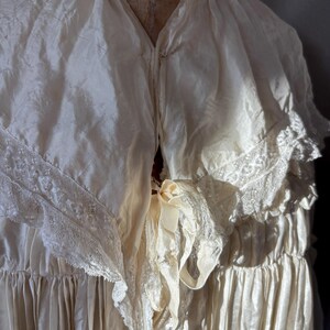 May include: Cream-colored vintage dress with a V-neckline and delicate lace trim. The dress features a flowing, layered design with a ribbon detail at the waist. The fabric appears to be silk or a similar lightweight material.